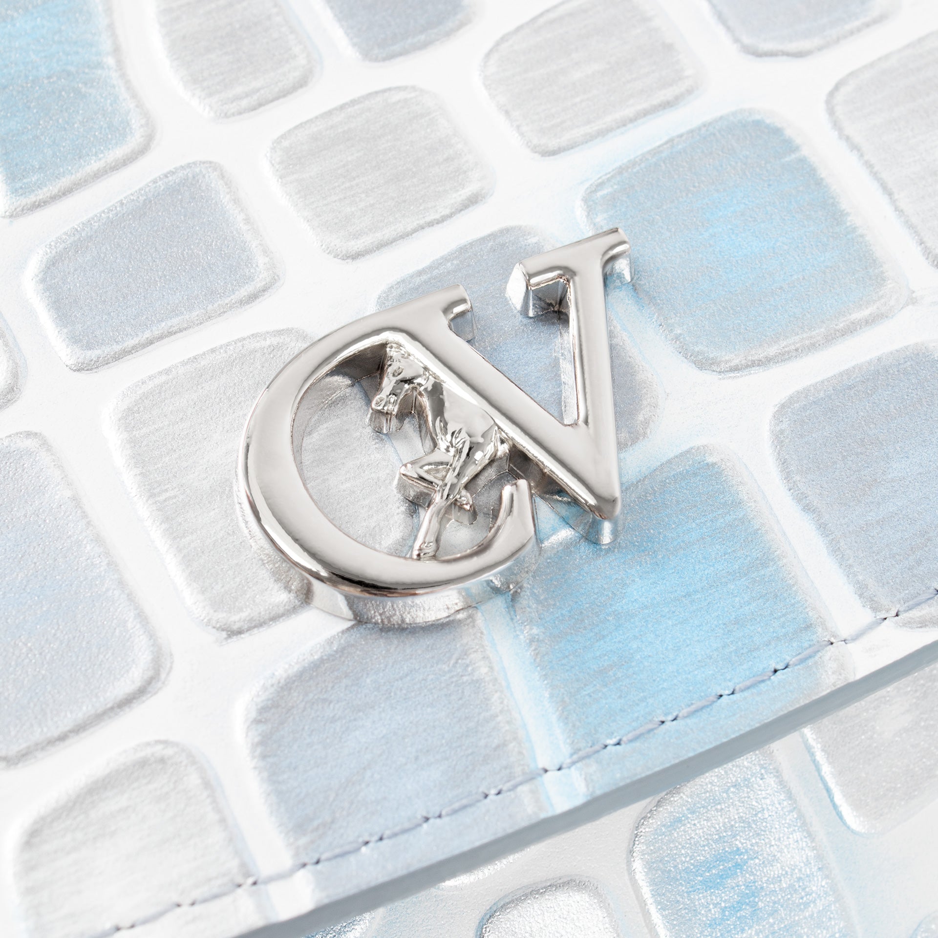 A close-up of a metallic silver interlocking 'CV' monogram charm with a small horse-and-carriage motif inside, resting on a textured white and pale blue surface.  #color_lightblue